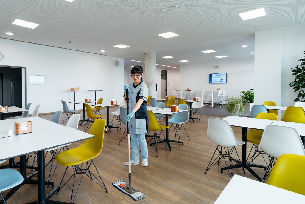 Mitarbeiterin von RWS Gebäudeservice wischt den Boden in einem modernen Aufenthaltsraum mit Tischen und Stühlen.